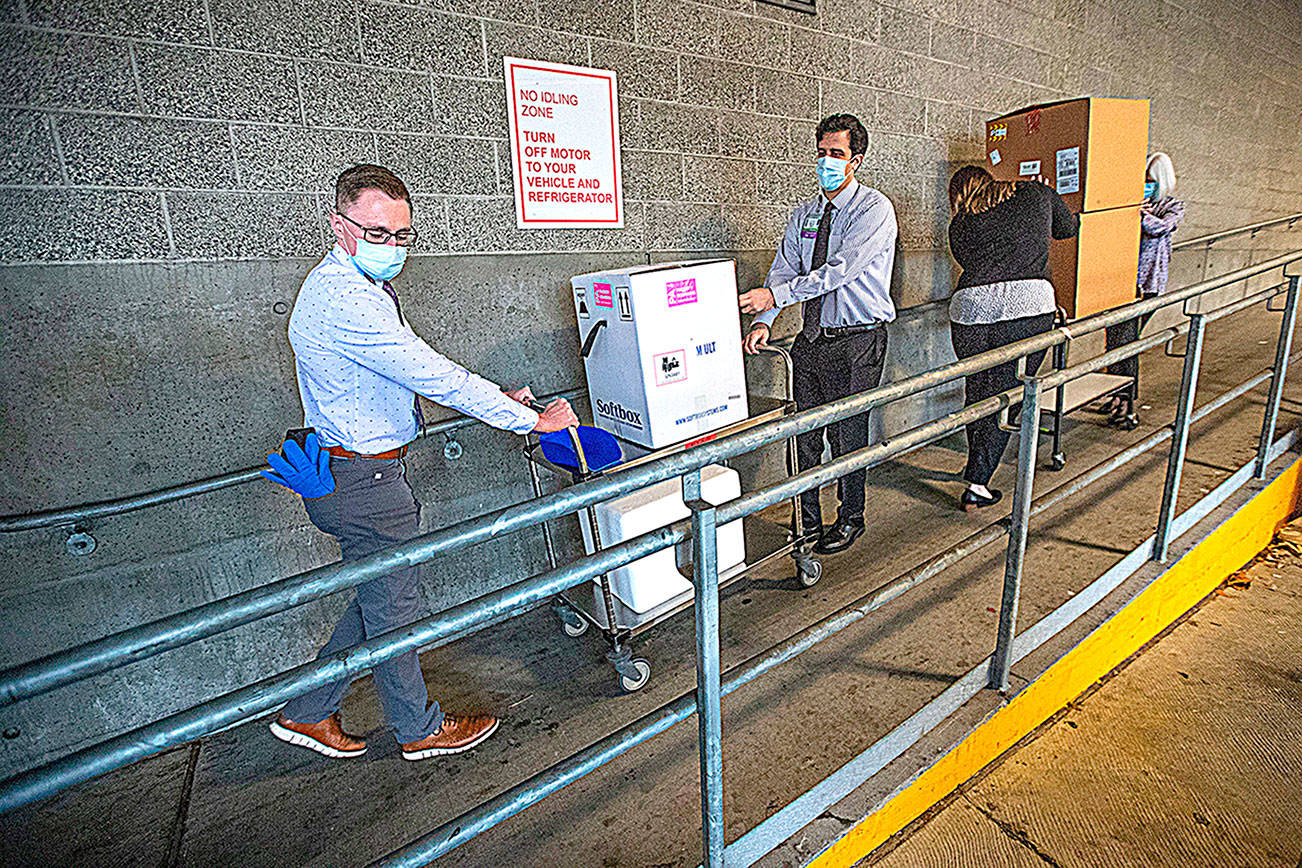 UWMC Montlake campus Pharmacy Administration Resident Derek Pohlmeyer, left, and UWMC Pharmacy Director Michael Alwan transport a box containing the Pfizer coronavirus vaccines toward a waiting vehicle headed to the UW Medicines other hospital campuses on Monday morning. These are among the first to be distributed in Washington state. (Pool Photo)