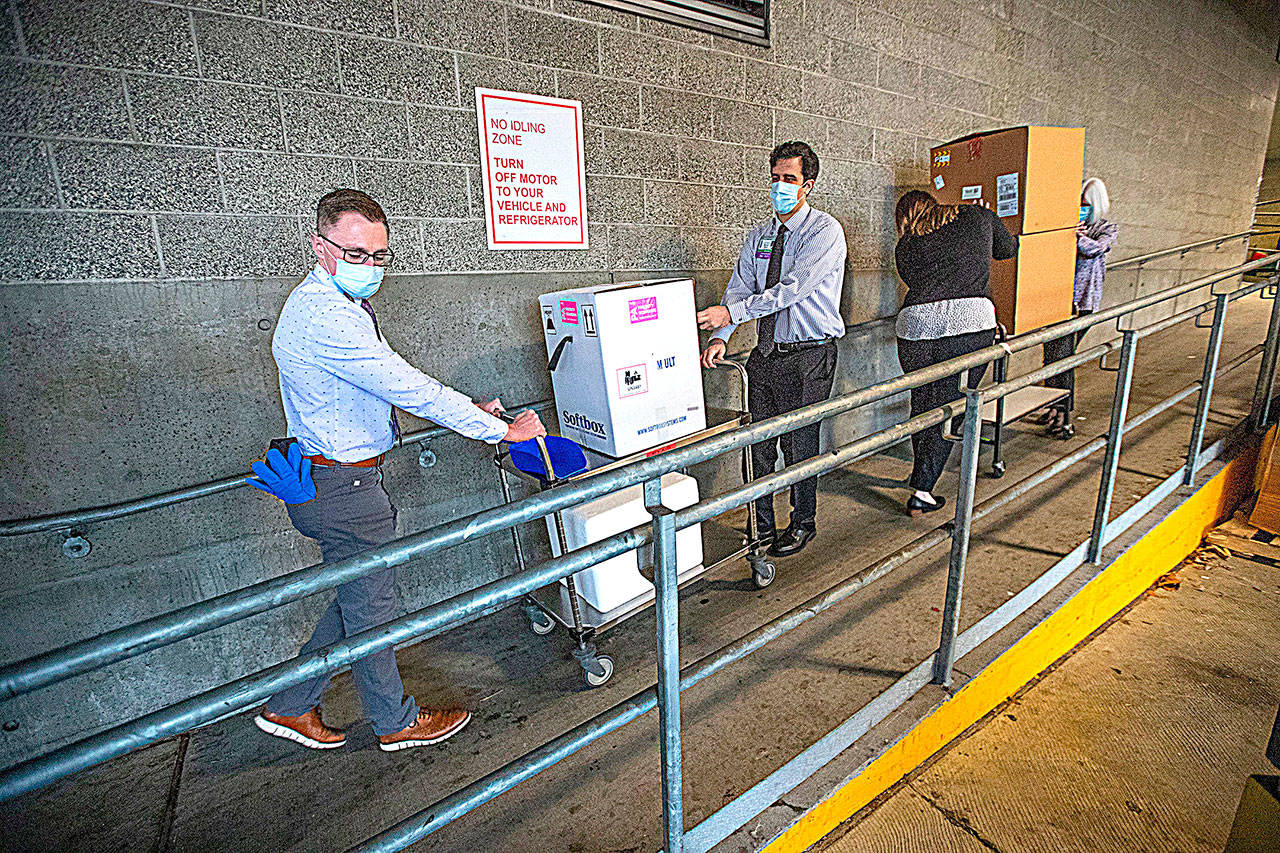 UWMC Montlake campus Pharmacy Administration Resident Derek Pohlmeyer, left, and UWMC Pharmacy Director Michael Alwan transport a box containing the Pfizer coronavirus vaccines toward a waiting vehicle headed to the UW Medicines other hospital campuses on Monday morning. These are among the first to be distributed in Washington state. (Pool Photo)